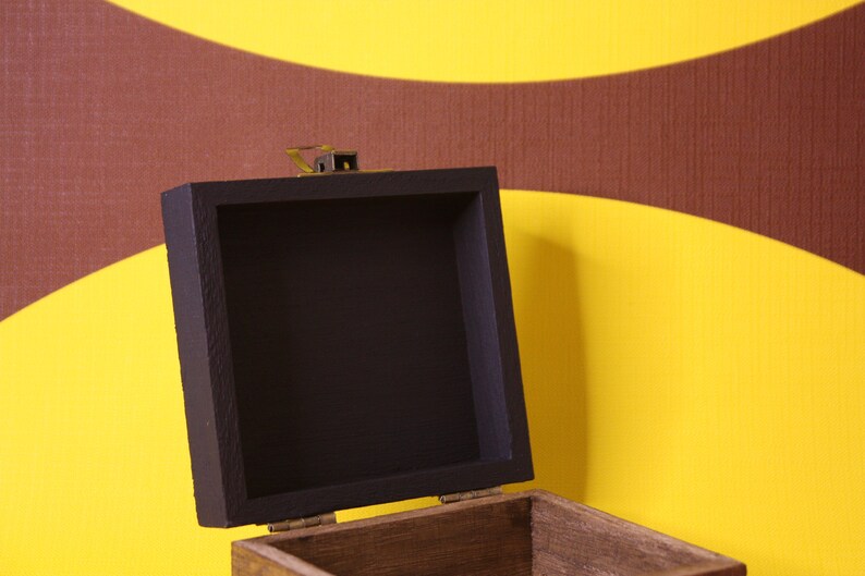 May include: An open wooden box with a black painted interior. The box has a brass latch and is sitting on a yellow and brown patterned background.