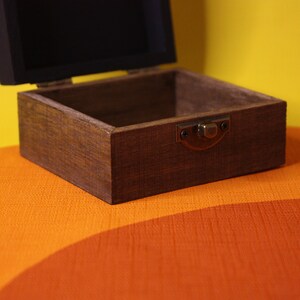 May include: A wooden box with a hinged lid and a brass latch. The box is open and empty, revealing a dark brown interior. The box is sitting on a multi-colored surface.