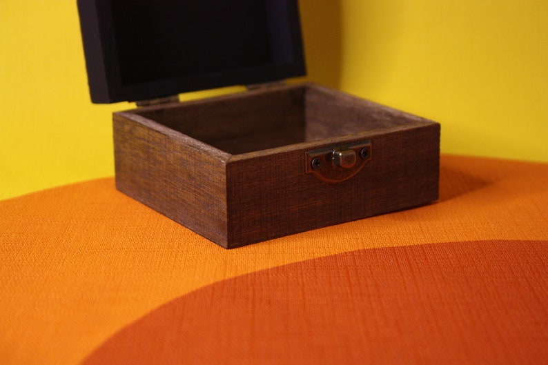 May include: A wooden box with a hinged lid and a brass latch. The box is open and empty, revealing a dark brown interior. The box is sitting on a multi-colored surface.