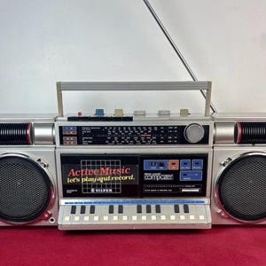 Op de afbeelding: Zilverkleurige boombox met een handvat, luidsprekers en een radioknop. De boombox heeft de tekst "Active Music let's play and record" en een klein toetsenbord. De boombox heeft een zilveren afwerking met zwarte en rode accenten.