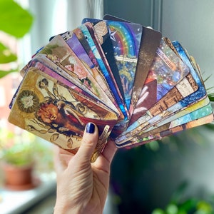 May include: A hand holding a fanned deck of tarot cards. The cards feature colorful illustrations with themes of nature, spirituality, and celestial elements. The cards have a vintage aesthetic, with gold and sepia tones. Some cards have text, including the word "manifestation".