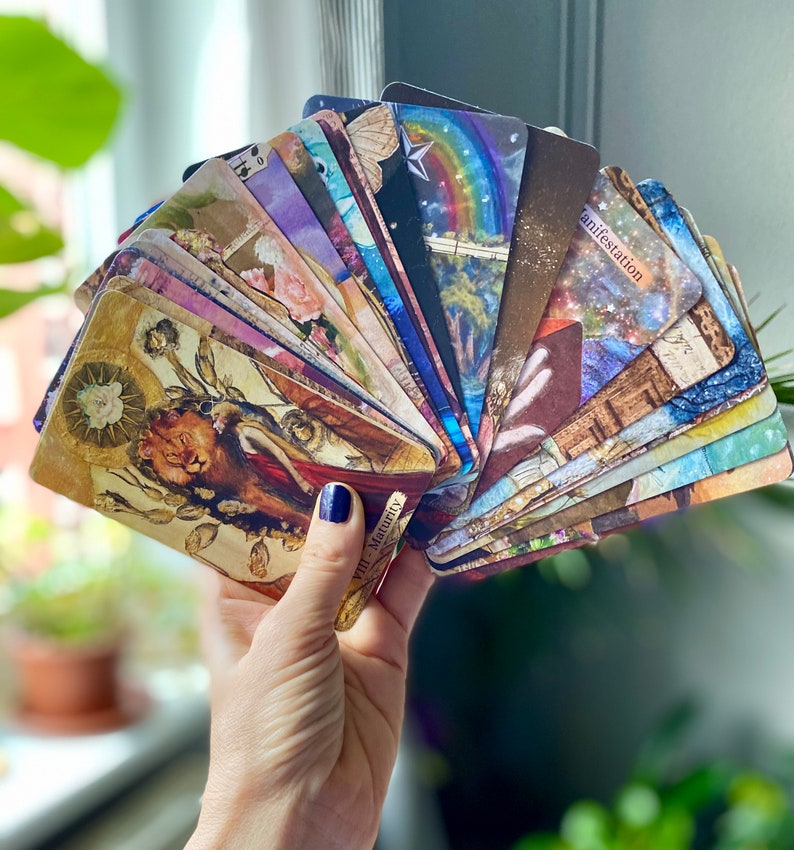 May include: A hand holding a fanned deck of tarot cards. The cards feature colorful illustrations with themes of nature, spirituality, and celestial elements. The cards have a vintage aesthetic, with gold and sepia tones. Some cards have text, including the word "manifestation".