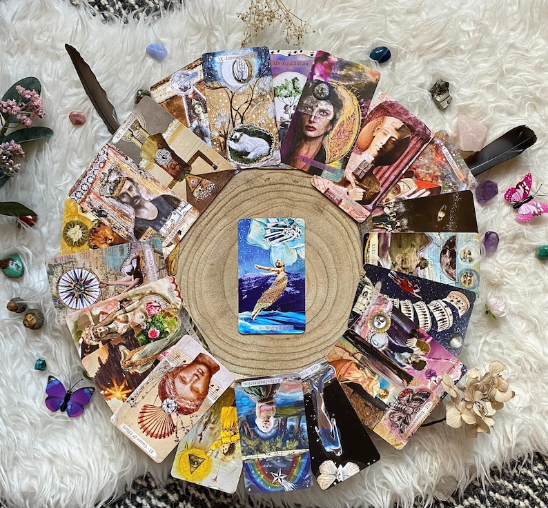 May include: A collection of tarot cards arranged in a semi-circle on a wooden slice, with a central card featuring a woman in a flowing dress. The cards display various illustrations and text, with decorative elements like feathers and crystals.