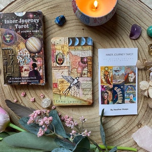 May include: A tarot card deck and guide book, titled "Inner Journey Tarot," are displayed on a wooden surface. The deck box features a planet graphic, while the cards have collage art. A lit candle, crystals, and flowers surround the deck.