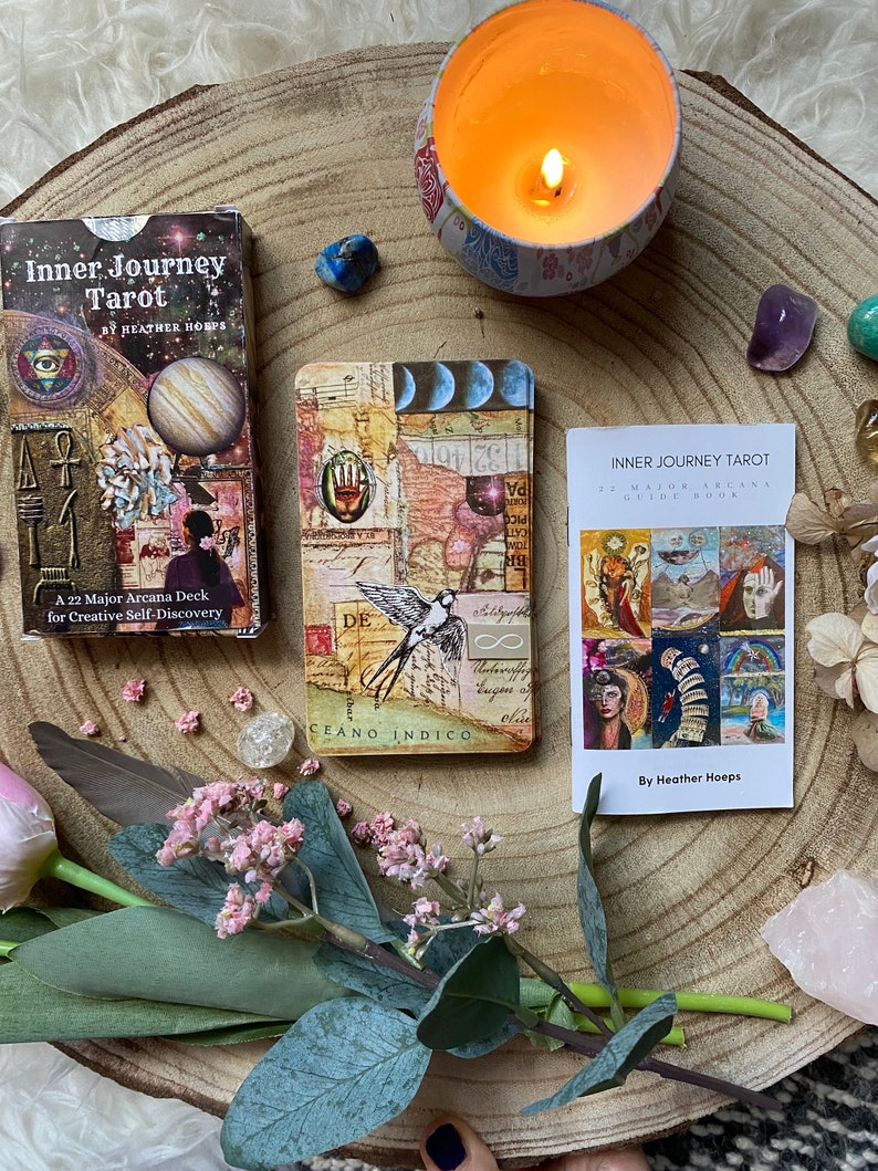 May include: A tarot card deck and guide book, titled "Inner Journey Tarot," are displayed on a wooden surface. The deck box features a planet graphic, while the cards have collage art. A lit candle, crystals, and flowers surround the deck.