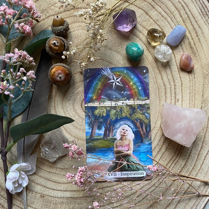 May include: A tarot card with a rainbow and star design, featuring a woman in a forest scene, surrounded by crystals, acorns, and flowers. The card reads "XVII - Inspiration".