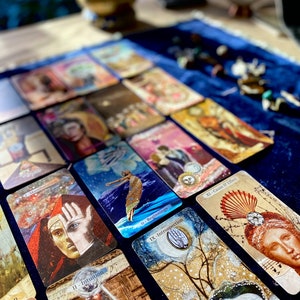 May include: A collection of tarot cards with various illustrations and text, including "0 - Beginnings", "II - Intuition", "IX - Introspection", and "XI - Cause & Effect". The cards are arranged on a dark blue velvet surface, with a blurred background featuring a brass bowl and other objects.