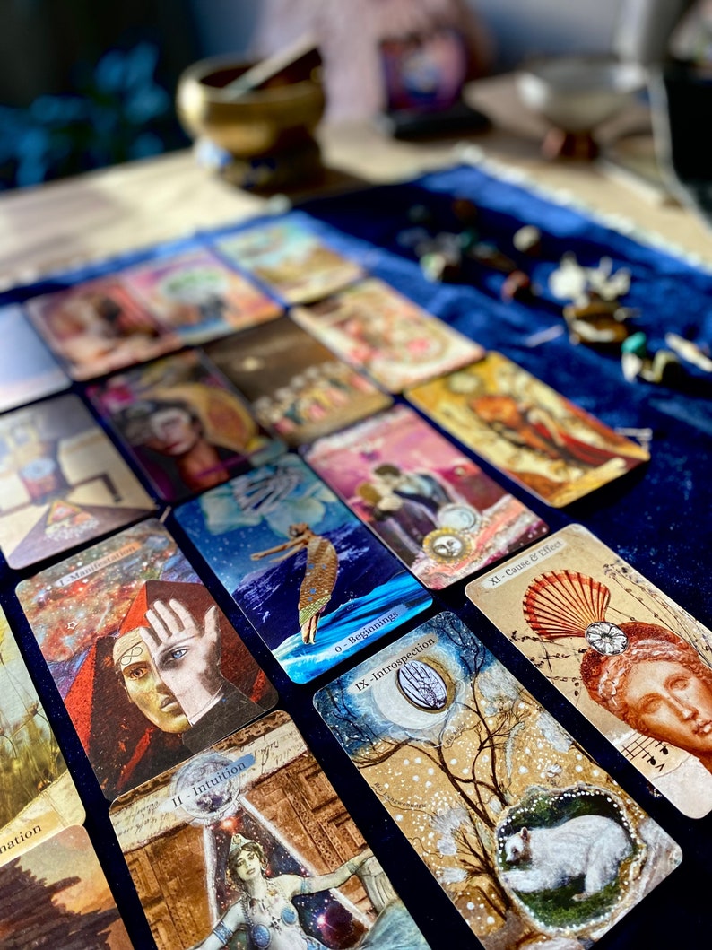 May include: A collection of tarot cards with various illustrations and text, including "0 - Beginnings", "II - Intuition", "IX - Introspection", and "XI - Cause & Effect". The cards are arranged on a dark blue velvet surface, with a blurred background featuring a brass bowl and other objects.