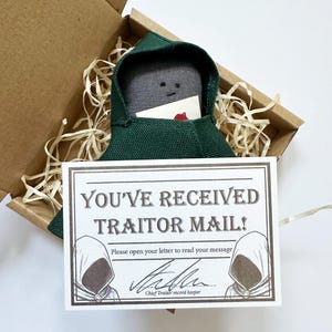 May include: A fabric doll in a green hooded cloak, presented in a cardboard box with straw-like packing. The doll holds a card with a red heart. A sign reads "YOU'VE RECEIVED TRAITOR MAIL!" with an illustration and signature.