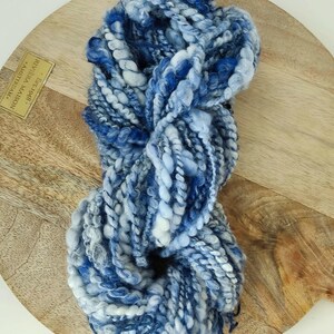 May include: A skein of thick, textured yarn featuring a blend of deep navy, royal blue, light blue, and white fibers. The yarn has a varied, chunky construction with twisted and looped strands, creating a multi-dimensional appearance.