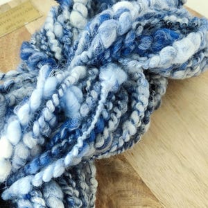 May include: A skein of thick, textured art yarn featuring a variegated color palette of deep navy, royal blue, sky blue, and white. The yarn consists of multiple plies with varying thicknesses, including smooth, fuzzy, and coiled sections.