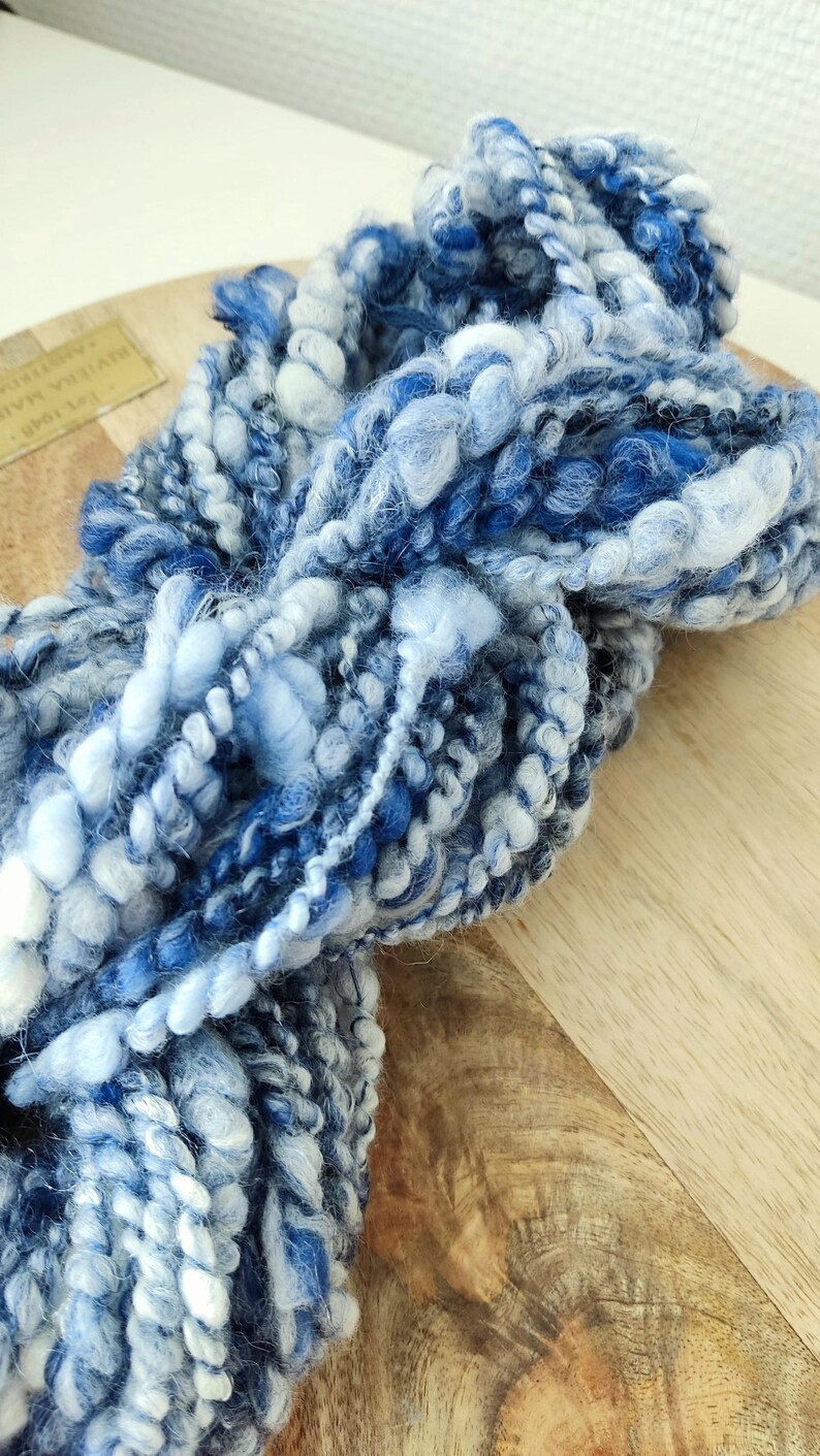 May include: A skein of thick, textured art yarn featuring a variegated color palette of deep navy, royal blue, sky blue, and white. The yarn consists of multiple plies with varying thicknesses, including smooth, fuzzy, and coiled sections.