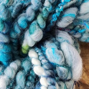 May include: A skein of thick, textured art yarn featuring a blend of blue, teal, turquoise, and white fibers. The yarn displays varying thicknesses, including chunky, nubby sections and smooth, twisted strands, creating a multi-dimensional appearance.