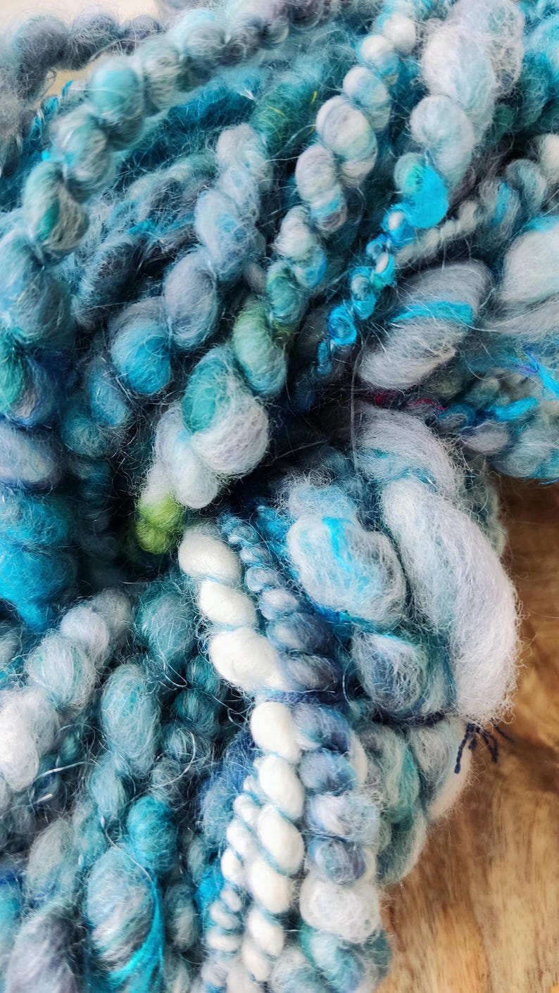 May include: A skein of thick, textured art yarn featuring a blend of blue, teal, turquoise, and white fibers. The yarn displays varying thicknesses, including chunky, nubby sections and smooth, twisted strands, creating a multi-dimensional appearance.