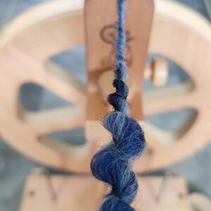 May include: A strand of dark blue and grey wool yarn being spun on a wooden spinning wheel. The yarn features a thick, textured twist as it feeds into the orifice of the spinning mechanism, with the blurred wooden wheel visible in the background.