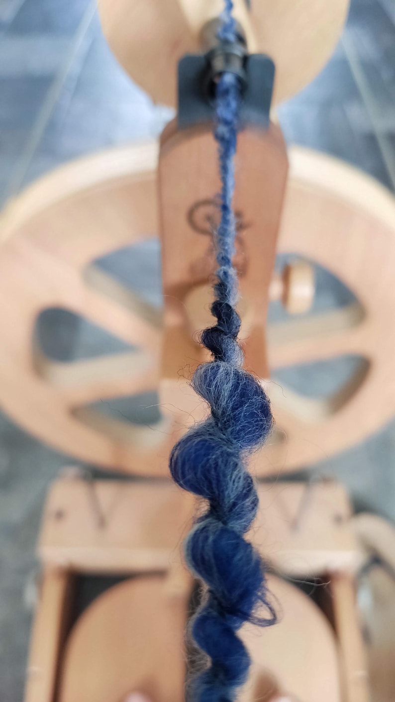 May include: A strand of dark blue and grey wool yarn being spun on a wooden spinning wheel. The yarn features a thick, textured twist as it feeds into the orifice of the spinning mechanism, with the blurred wooden wheel visible in the background.