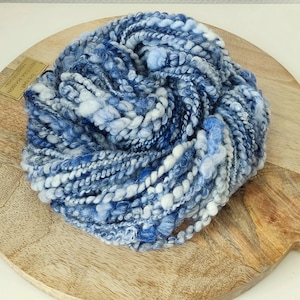 May include: A skein of thick, textured art yarn featuring a variegated color palette of deep navy, royal blue, sky blue, and white. The yarn consists of multiple plies with varying thicknesses, including smooth, fuzzy, and nubby sections.