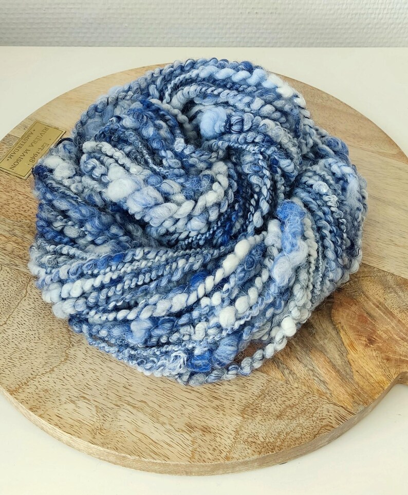 May include: A skein of thick, textured art yarn featuring a variegated color palette of deep navy, royal blue, sky blue, and white. The yarn consists of multiple plies with varying thicknesses, including smooth, fuzzy, and nubby sections.
