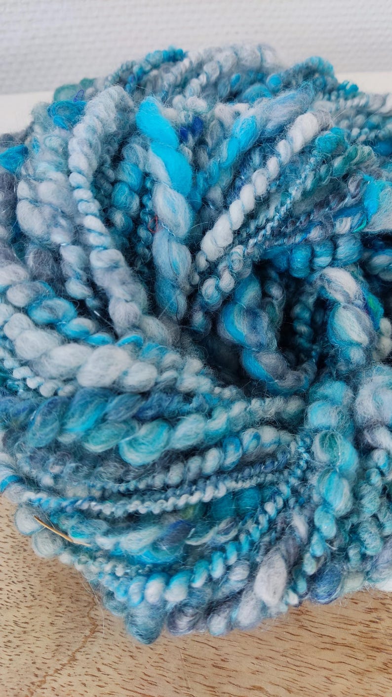 May include: A ball of art yarn featuring a blend of blue, turquoise, light blue, and grey fibers. The yarn has a highly textured, uneven appearance with varying thicknesses, including thick, fluffy sections and thinner, tightly twisted strands.