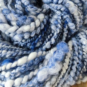May include: A skein of hand-spun art yarn featuring a blend of various blue and white fibers. The yarn displays a highly textured, irregular construction with thick, coiled, and fuzzy sections, creating a multi-dimensional appearance throughout.