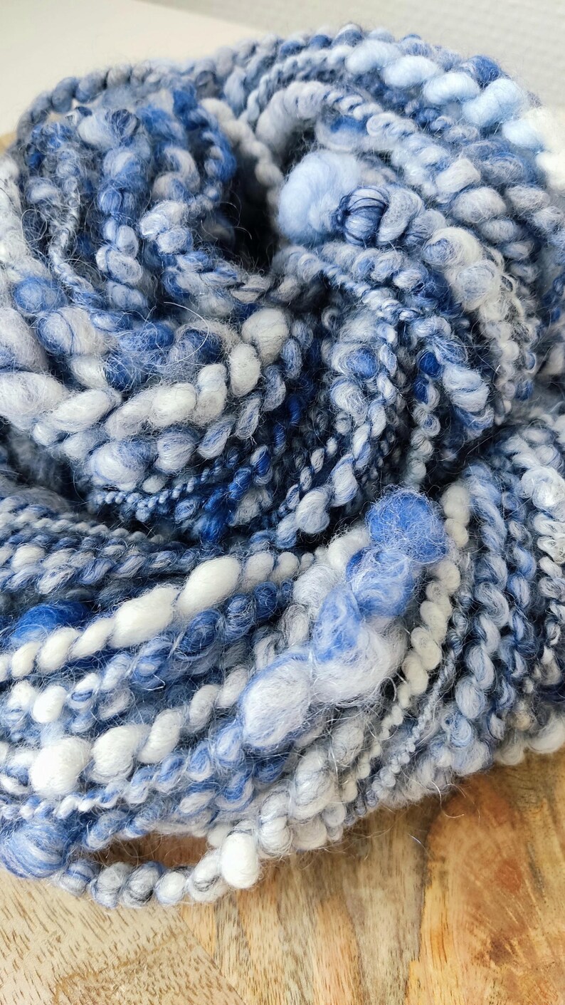 May include: A skein of hand-spun art yarn featuring a blend of various blue and white fibers. The yarn displays a highly textured, irregular construction with thick, coiled, and fuzzy sections, creating a multi-dimensional appearance throughout.