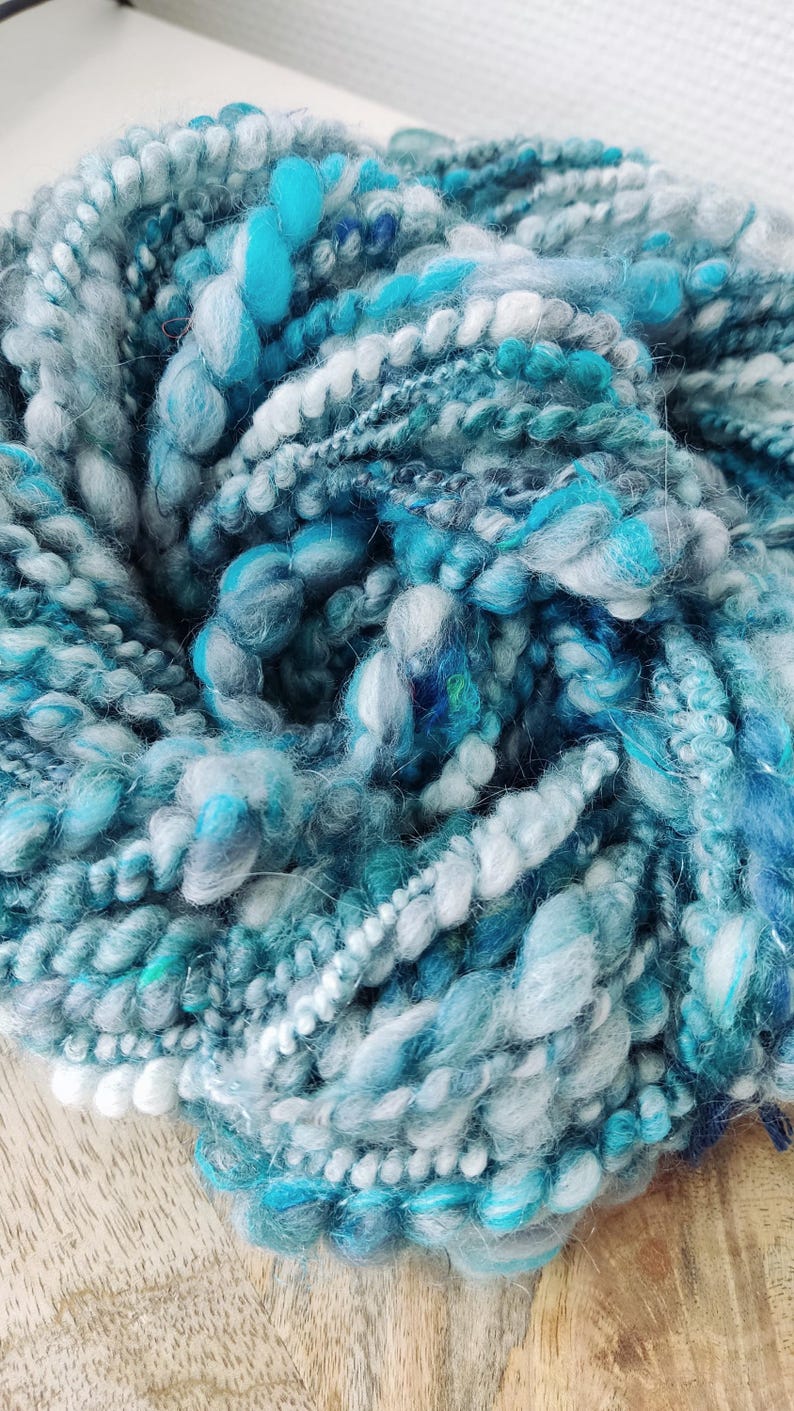 May include: A skein of hand-spun art yarn featuring a textured blend of fibers in shades of turquoise, teal, sky blue, navy, and white. The yarn displays varying thicknesses, including thick, fluffy sections and thinner, tightly twisted strands.