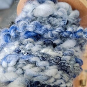 May include: A thick, hand-spun yarn wrapped around a wooden bobbin. The yarn features a variegated color palette of deep navy blue, bright royal blue, light sky blue, and crisp white, with a textured, uneven ply and soft, fuzzy fibers.