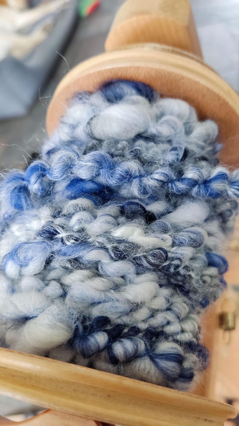 May include: A thick, hand-spun yarn wrapped around a wooden bobbin. The yarn features a variegated color palette of deep navy blue, bright royal blue, light sky blue, and crisp white, with a textured, uneven ply and soft, fuzzy fibers.