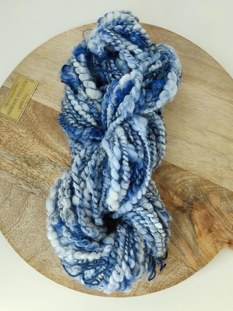May include: A skein of thick, textured yarn featuring a blend of deep navy, royal blue, light blue, and white fibers. The yarn has a varied, chunky construction with twisted and looped strands, creating a multi-dimensional appearance.