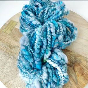 May include: A skein of hand-spun art yarn featuring a variegated color palette of deep ocean blue, turquoise, sky blue, and white. The yarn has a highly textured, irregular construction with thick and thin sections, loops, and twisted plies.