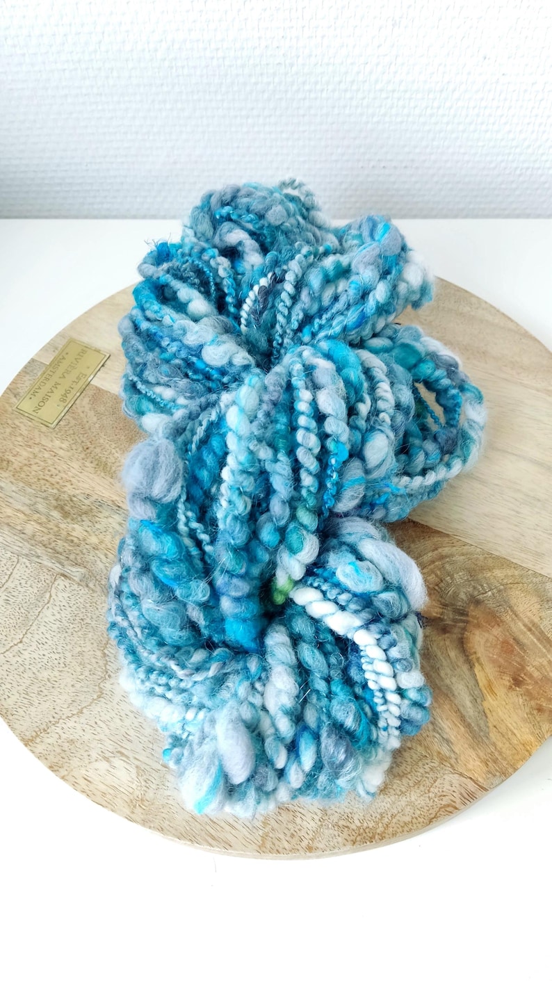 May include: A skein of hand-spun art yarn featuring a variegated color palette of deep ocean blue, turquoise, sky blue, and white. The yarn has a highly textured, irregular construction with thick and thin sections, loops, and twisted plies.