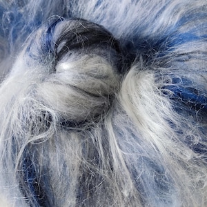 May include: A ball of fluffy yarn featuring a blend of white, light blue, and dark navy blue fibers. The texture is soft and fibrous, with long, loose strands creating a multi-tonal appearance throughout the round, coiled structure of the yarn.