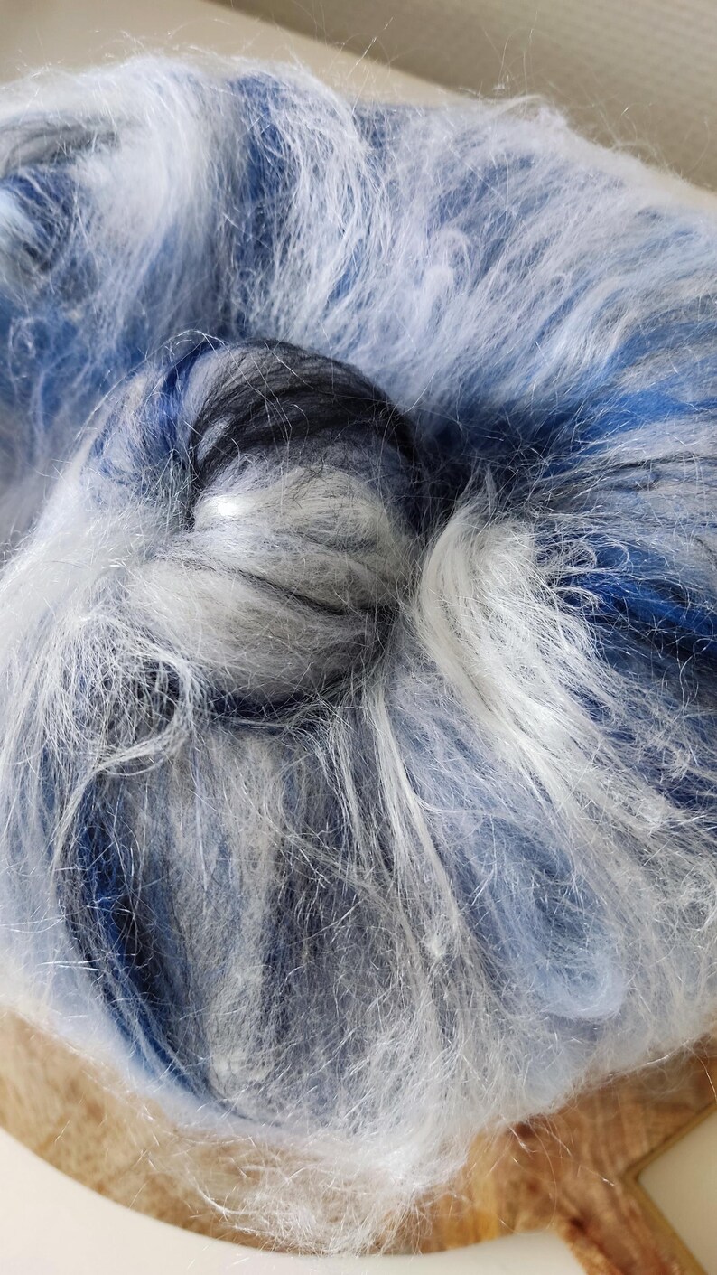 May include: A ball of fluffy yarn featuring a blend of white, light blue, and dark navy blue fibers. The texture is soft and fibrous, with long, loose strands creating a multi-tonal appearance throughout the round, coiled structure of the yarn.