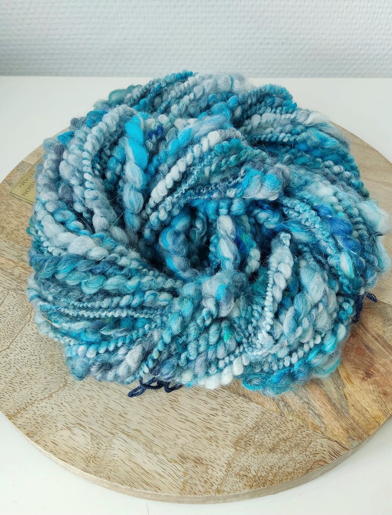 May include: A circular skein of hand-spun art yarn featuring a variegated color palette of deep navy, bright turquoise, sky blue, and soft white. The yarn displays a highly textured, thick-and-thin construction with varied ply twists.