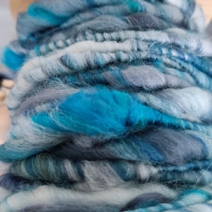 May include: A wooden spinning wheel bobbin filled with hand-spun yarn featuring a variegated color palette of bright turquoise, slate gray, charcoal, and white. The yarn has a thick, textured, and uneven ply with visible fiber tufts throughout.