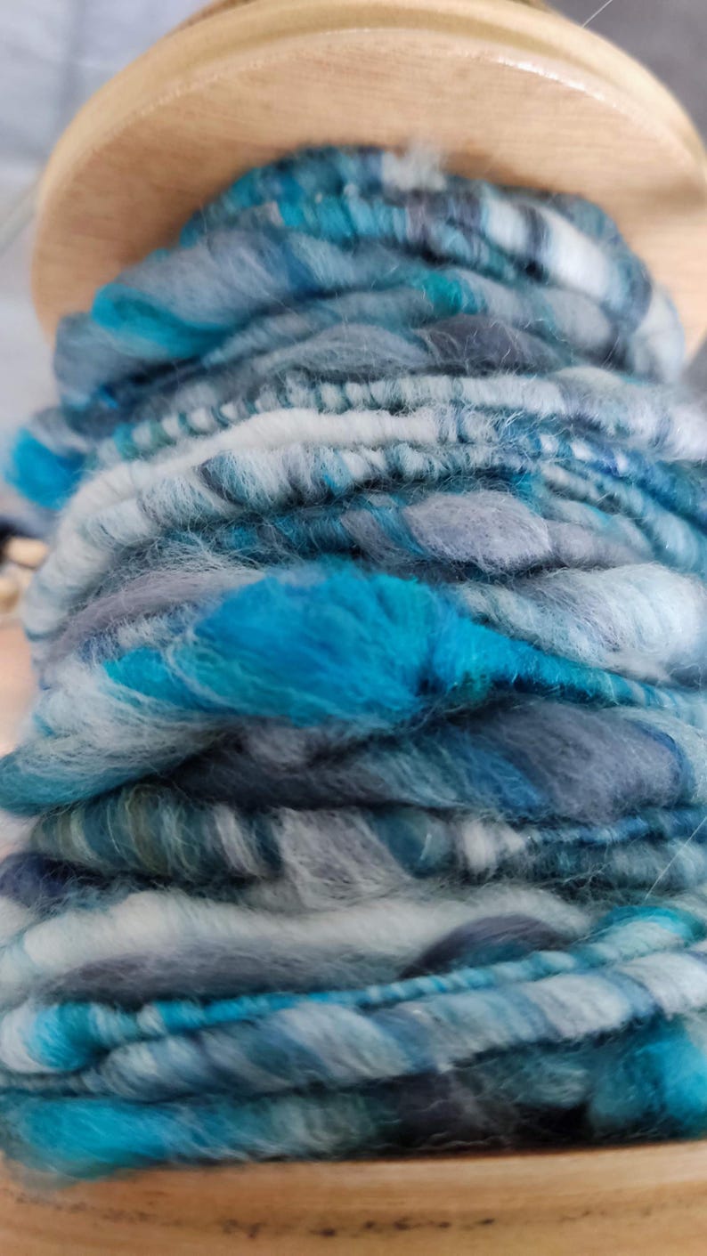 May include: A wooden spinning wheel bobbin filled with hand-spun yarn featuring a variegated color palette of bright turquoise, slate gray, charcoal, and white. The yarn has a thick, textured, and uneven ply with visible fiber tufts throughout.