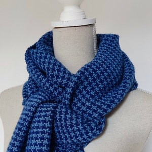 Op de afbeelding: Blauwe sjaal met een herhalend bloemmotief, gedrapeerd over een mannequin. De sjaal is een diepe blauwtint met een lichter blauw patroon. Gemaakt van zacht, getextureerd materiaal, ideaal voor koud weer.