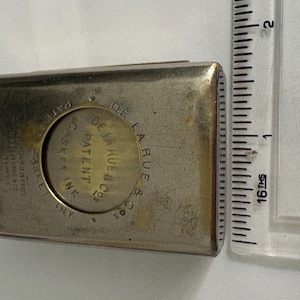 May include: A rectangular silver-toned metal ink casket case measuring approximately 5.7 cm in length. The front features a circular embossed design with the text 'PATENT DE LA RUE & CO CASKET INK' and additional small text on the left side.
