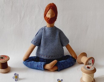 May include: A handmade fabric doll with a red beard and blue and white striped shirt sits in a meditative pose on a white surface with two spools of thread and blue flowers.
