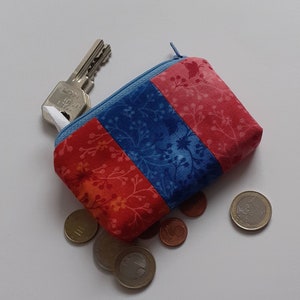 Peut inclure: Une petite bourse à monnaie en tissu rouge et bleu avec une fermeture éclair. La bourse est ouverte et des pièces d'euro en tombent.