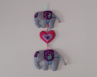 May include: A gray felt elephant with purple and pink details hangs from a white wall. A pink heart with a beaded border hangs between two elephants.