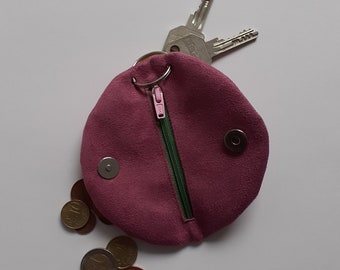 May include: A small, round, pink suede coin purse with a green zipper and a silver key ring attached. The purse is open and coins are spilling out.