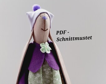 May include: A purple and green fabric doll with long ears, a purple dress, and a white hat. The doll is standing on a white surface with spools of thread and small blue flowers.