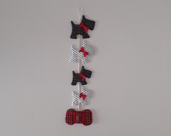May include: A string of five black and white fabric Scottie dog ornaments with red bows hanging from a white ceiling. The bottom ornament is a red and black plaid bow.