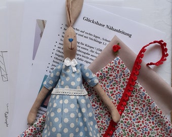 May include: A blue and white polka dot dress with a white lace trim and a white bow. The dress is on a stuffed bunny with brown ears and a white bow. The bunny is on a fabric with red and white flowers. The text "Glückshase Nähanleitung" is visible on a white piece of paper.