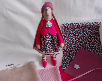 May include: A pink fabric bunny doll with a pink dress, a pink jacket, and a pink hat. The doll is surrounded by fabric scraps in various colors and patterns, including pink, red, and blue.