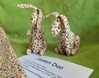 May include: Two white fabric bunnies with a floral pattern in shades of pink, blue, and green. The bunnies are sitting on a green surface. The text "Hasen Duo" is written on a white piece of paper in front of the bunnies.