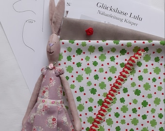 May include: A pink fabric bunny doll with a floral print dress and a white ribbon around the waist. The doll is sitting on a white fabric with a green and red clover pattern.