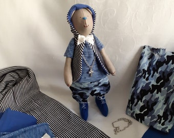 May include: A handmade plush toy bear wearing a blue hooded shirt, a striped vest, blue camouflage pants, and blue shoes. The bear has a white bow tie and a silver chain with a cross pendant. The toy is surrounded by fabric scraps in various shades of blue.