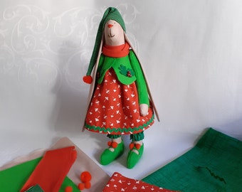 May include: A handmade plush toy rabbit wearing a green and red dress with a white mushroom pattern. The rabbit has a green hat with white stripes and a red pom-pom. The toy is surrounded by fabric scraps in green and red.